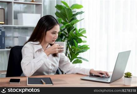 Young asian enthusiastic businesswoman at modern office desk using laptop to work with a cup of coffee. Diligent and attractive office lady working on computer notebook in her office work space.. Young asian enthusiastic businesswoman at modern office desk with laptop.