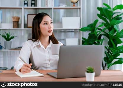 Young asian enthusiastic businesswoman at modern office desk using laptop to work with a cup of coffee. Diligent and attractive office lady working on computer notebook in her office work space.. Young asian enthusiastic businesswoman at modern office desk with laptop.