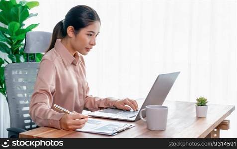 Young asian enthusiastic businesswoman at modern office desk using laptop to work and write notes. Diligent and attractive office lady working on computer notebook in her office work space.. Young asian enthusiastic businesswoman at modern office desk with laptop.