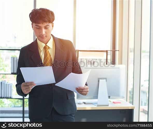 Young asian experienced entrepreneur in formal wear reading financial report and calling to manager on smartphone to discuss data.Businessman with paper document in hands talking on cellular in office