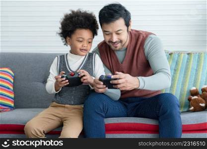 Young Asian father with beard and African little kid son enjoy playing games holding joystick controller together sitting on sofa at living room at home. Relationship of diverse family