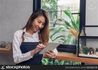 Young Asian female barista using electronic tablet in her own coffee shop while takeing break in warm afternoon. Young female barista and her modern small coffee shop. Food and drink industry concept.