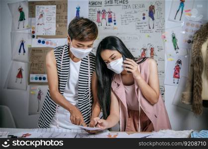 Young asian female designer and colleague wearing medical mask during working in modern tailor shop, distance and new normal concept 