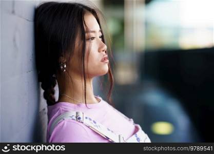 Young Asian female in casual clothes with braid leaning on wall and looking away on city street in daytime. Asian woman leaning on wall