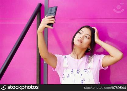 Young Asian female in stylish clothes touching head and taking selfie against pink wall of modern building. Asian woman taking selfie against pink wall