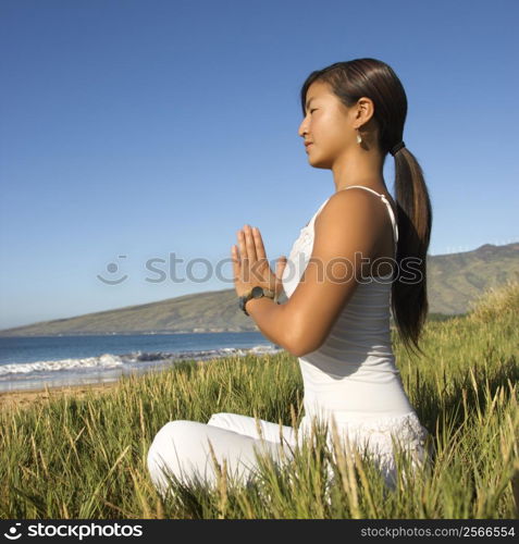 Young Asian female sitting on beach looking out to ocean with hands pressed together.