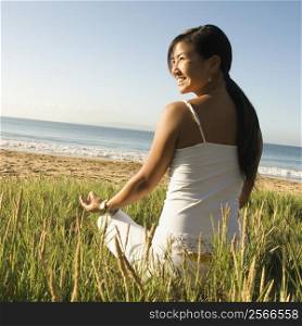 Young Asian female sitting on beach meditating and looking off to the side.