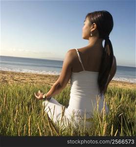 Young Asian female sitting on beach meditating and looking out to ocean.