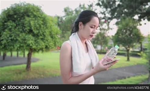 Young asian female using mobile phone while exercising at the park, holding plastic water bottle, enjoy exercise body care, pure fresh water, wireless online connection, enjoyment positive emotion