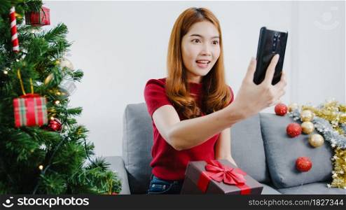 Young Asian female using smart phone video call talking with couple with X&rsquo;Mas present box, Christmas tree decorated with ornament in living room at home. Christmas and New Year holiday festival.
