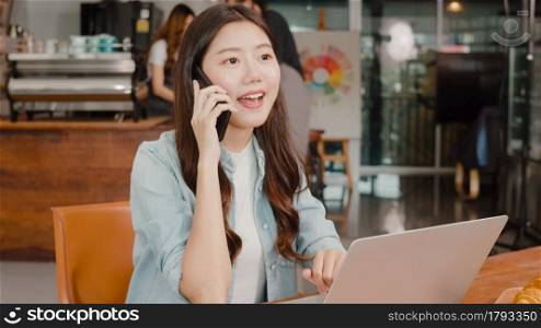 Young Asian freelance female work talk on phone with customer at coffee shop. Attractive Japanese business woman is making a business call during the lunch break in cafe or restaurant with laptop.
