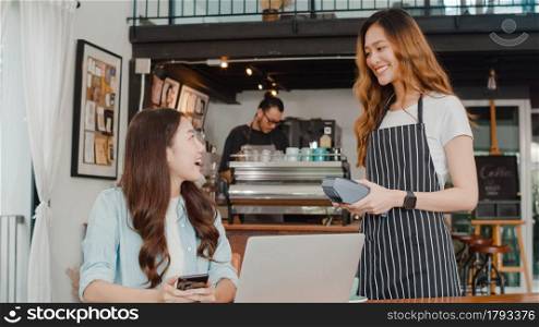 Young Asian freelance women pay contactless at coffee shop. Asian happy girl barista waiter wear gray apron holding credit card reader machine for customer using mobile phone scan pay in cafe.