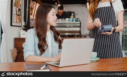 Young Asian freelance women pay contactless at coffee shop. Asian happy female barista waiter wear gray apron holding credit card reader machine for customer using mobile phone scan pay in cafe.