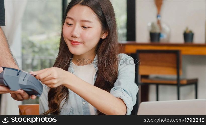Young Asian freelance women pay contactless credit card at coffee shop. Asian happy men barista waiter wear gray apon holding credit card reader machine for customer can pay with technology in cafe.
