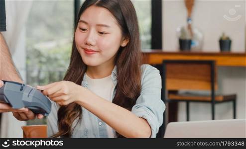 Young Asian freelance women pay contactless credit card at coffee shop. Asian happy men barista waiter wear gray apon holding credit card reader machine for customer can pay with technology in cafe.