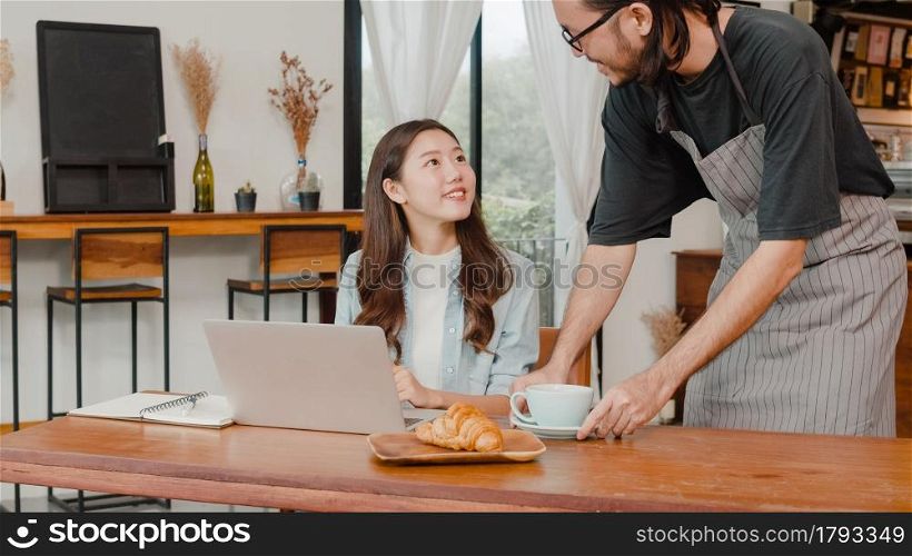 Young Asian freelance women work talk on phone with customer at coffee shop. Asian happy men barista waiter wear gray apon serve hot coffee to female customer who business work in cafe or restaurant.
