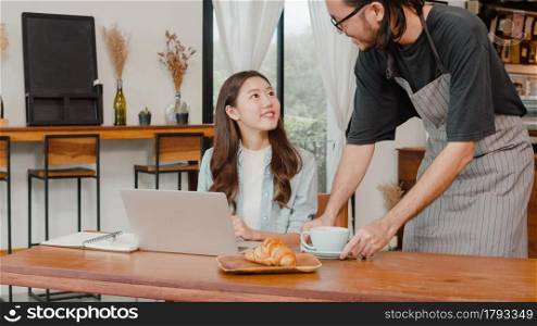 Young Asian freelance women work talk on phone with customer at coffee shop. Asian happy men barista waiter wear gray apon serve hot coffee to female customer who business work in cafe or restaurant.