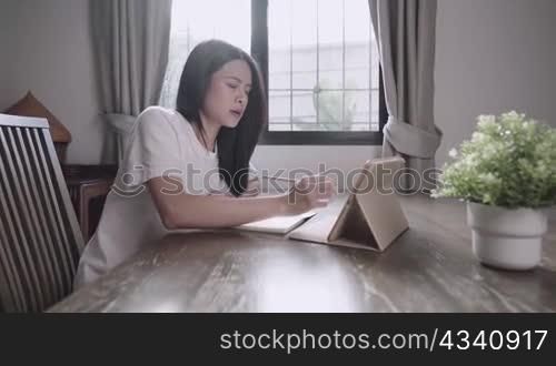 Young asian girl working on final exam revision, using tablet for online class lesson, at home schooling, use pencil write down on small notebook, job searching opportunity, sitdown alone at home desk