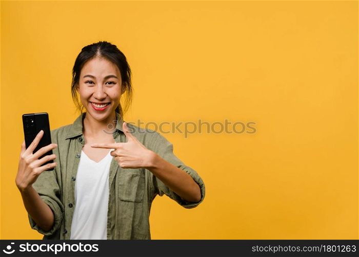 Young Asian lady using mobile phone with cheerful expression, shows something amazing at blank space in casual cloth and looking at camera isolated over yellow background. Facial expression concept.