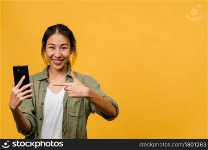 Young Asian lady using mobile phone with cheerful expression, shows something amazing at blank space in casual cloth and looking at camera isolated over yellow background. Facial expression concept.