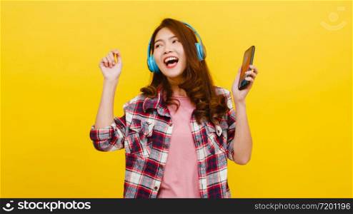 Young Asian lady wearing wireless headphones listening to music from smartphone with cheerful expression in casual clothing and looking at camera over yellow background. Facial expression concept.