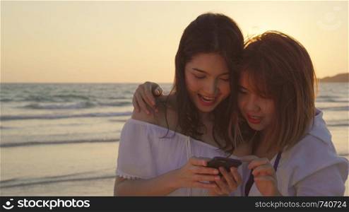 Young Asian lesbian couple using smart phone near beach. Beautiful women lgbt couple happy relax enjoy love moment when sunset in evening. Lifestyle lesbian couple travel on beach concept