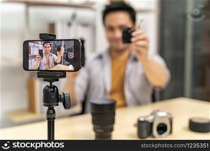 Young asian male IT vlogger and blogger live about digital camera Technology upgrand using mobile phone to recording live vlog video. Online influcencer on social media concept. Focus on camera.