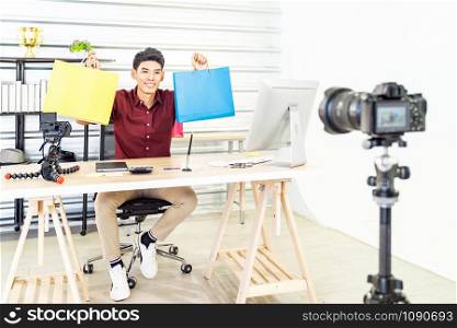 Young asian male online merchant blogger recording live vlog video with colorful shopping bag preparing for sale and review goods at home. Online influcencer on social media concept.