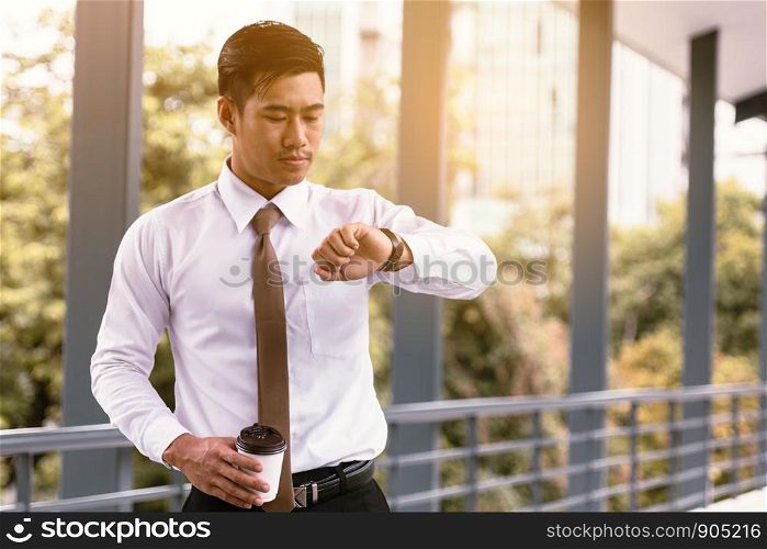 Young asian male standing checking time and holding coffee cup with confident looking at watches on hand at outside company.
