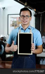 Young asian man barista holding blank chalkboard and thumb up with smiling face at cafe counter background, food and drink concept