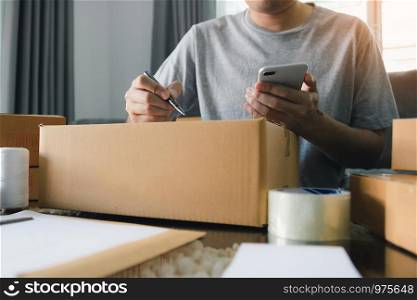 Young asian man business owner hands writing address on cardboard box at workplace or home office.