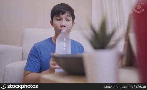 Young Asian man drinking water from big plastic bottle simple lifestyle inside the house, stay hydrate, sit down on the floor sofa on the background, hungry man eating and enjoy his home cook dinner