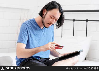 Young asian man holding credit card and using digital tablet for shopping on line at home, business and technology concept, digital marketing, casual lifestyle