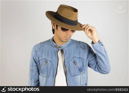 Young Asian Man in Jeans Jacket or Denim Jacket Wear Sunglasses and Touch Hat Brim in 45 Degree Pose. Denim or Jeans Jacket Men Fashion on Gray Background