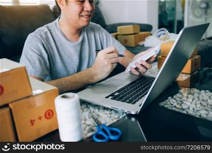 Young asian man looking on computer laptop and writing order list on paper, SME concept.