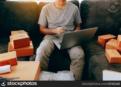 Young asian man sitting on sofa looking on computer laptop and writing order list on paper, SME concept.