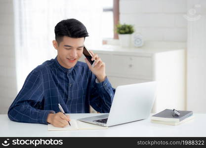 Young asian man talking phone and work from home with laptop computer, freelance male writing on notebook and speak on smartphone, stay home, domestic life, business and communication concept.