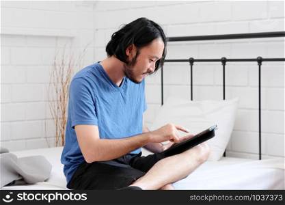 Young asian man using tablet while sitting on bed in white room background, people and technology, lifestyle, adult online education, working from home
