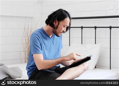 Young asian man using tablet while sitting on bed in white room background, people and technology, lifestyle, adult online education, working from home