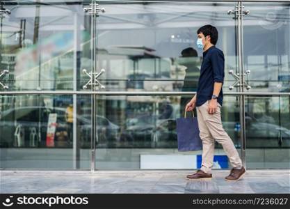 young asian man walking and holding shopping bag in front of store and his wearing medical mask for prevention from coronavirus (Covid-19) pandemic. new normal concepts