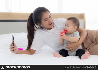 Young asian mother and little baby girl or newborn selfie with smart phone on bed in bedroom, happiness mom and daughter using phone video call at home, two people, family and communication concept.