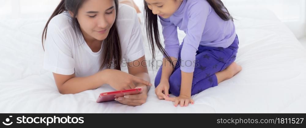 Young asian mother and little daughter looking tablet computer together for learning and education on bed in bedroom, mom and girl watching tablet to internet at home, family and technology concept.