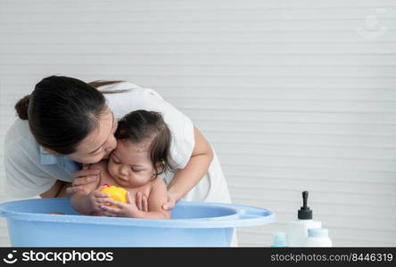 Young Asian mother bathing her little baby at home and kissing child head while cute daughter enjoy playing yellow duck toy and sitting in bathtub, Baby bathing concept. White background