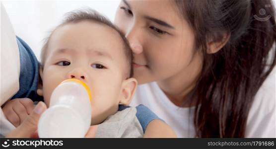 Young asian mother embracing and feeding little baby girl with bottle of milk at home, newborn innocence drinking with mom satisfied, relationship and bonding of mum and child, family concept.