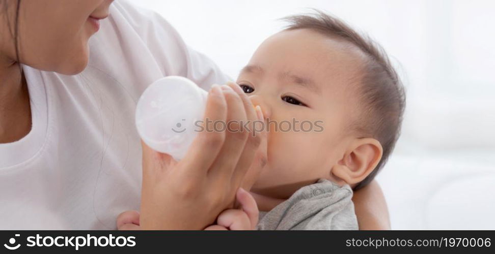 Young asian mother embracing and feeding little baby girl with bottle of milk at home, newborn innocence drinking with mom satisfied, relationship and bonding of mum and child, family concept.