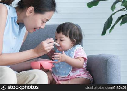 Young Asian mother holding spoon feeding child, Caucasian Cute 7 month newborn baby girl eating blend mashed food, sitting on sofa with apron at home, baby mouth mess up with food. Healhy food for kid