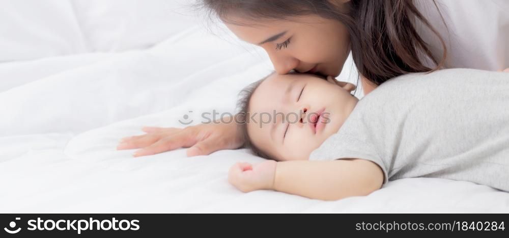 Young asian mother kiss cheek of little baby girl with tender on bed in the bedroom, mom love newborn and care, mother with expression with child together, parent and daughter, family concept.