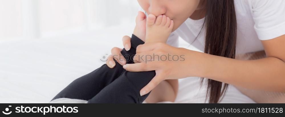 Young asian mother kiss foot of little baby girl with tender on bed in the bedroom, mom love newborn and care, mother with expression with child together, parent and daughter, family concept.