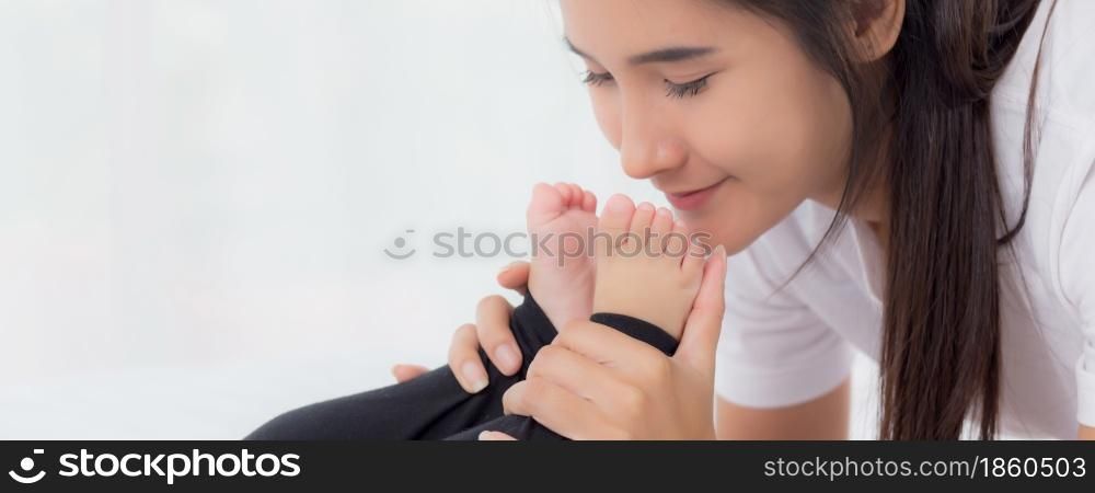 Young asian mother kiss foot of little baby girl with tender on bed in the bedroom, mom love newborn and care, mother with expression with child together, parent and daughter, family concept.