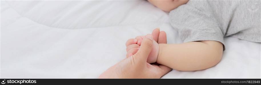 Young asian mother touch hand of little baby girl with tender on bed in the bedroom, mom love newborn and care, woman with expression with child together, parent and daughter, family concept.